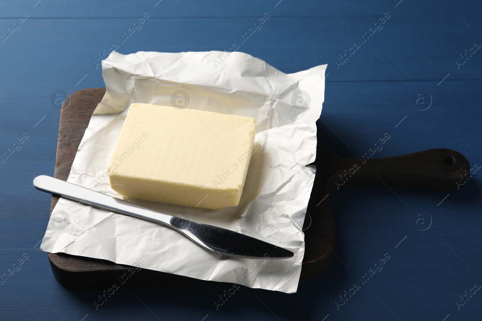 Photo of Block of tasty butter in open foil packaging and knife on blue wooden table