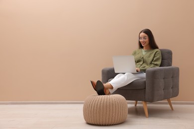 Beautiful woman with laptop sitting in armchair near beige wall indoors, space for text