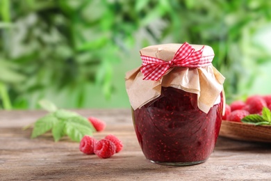 Photo of Glass jar of sweet jam with ripe raspberries on wooden table against blurred background. Space for text