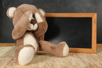 Teddy bear covering eyes and small blackboard on wooden table. Space for text