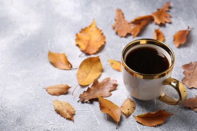 Cup of hot drink and autumn leaves on light grey textured table, space for text