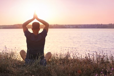 Man practicing yoga near river at sunset, back view