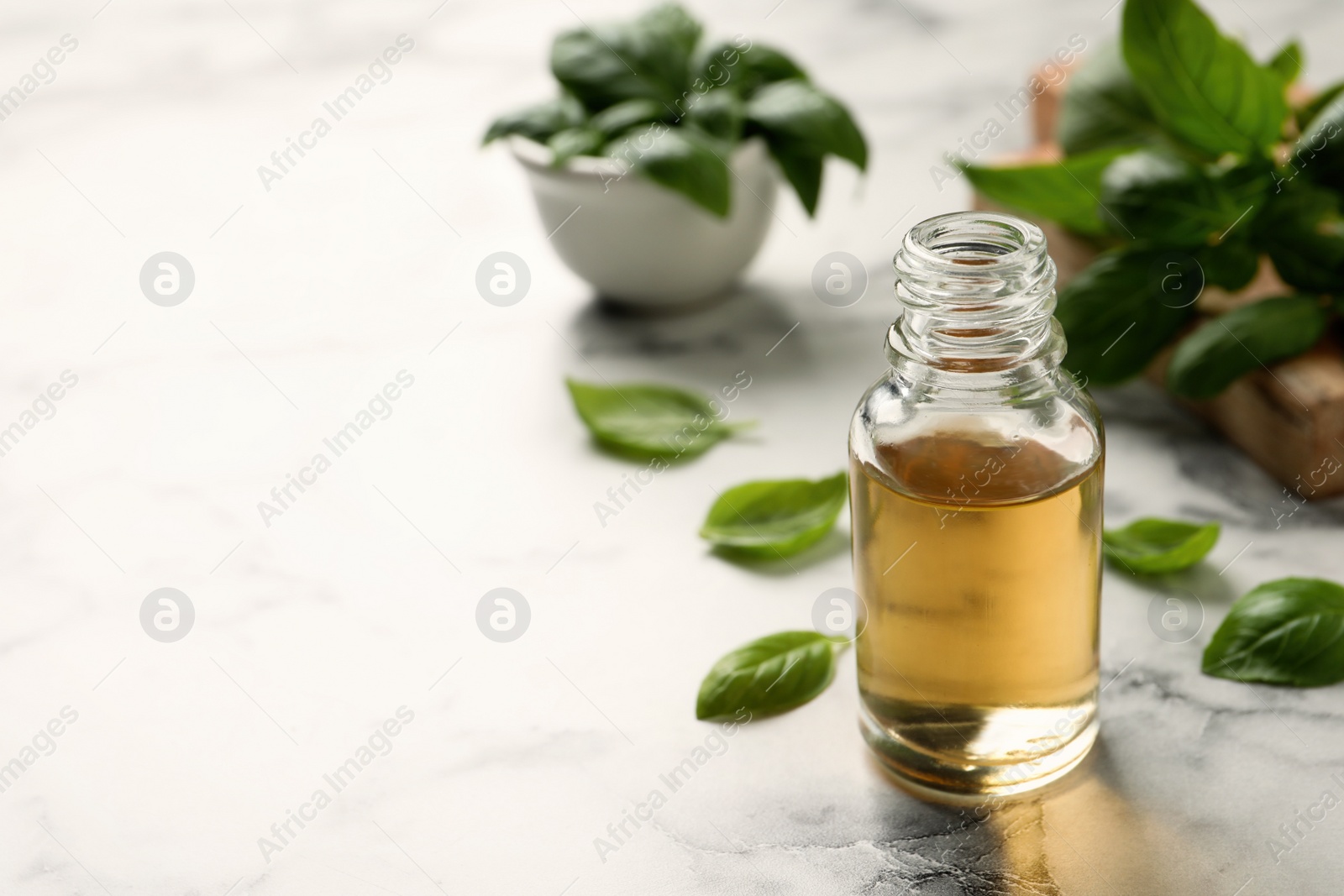 Photo of Glass bottle of basil essential oil and leaves on white marble table. Space for text