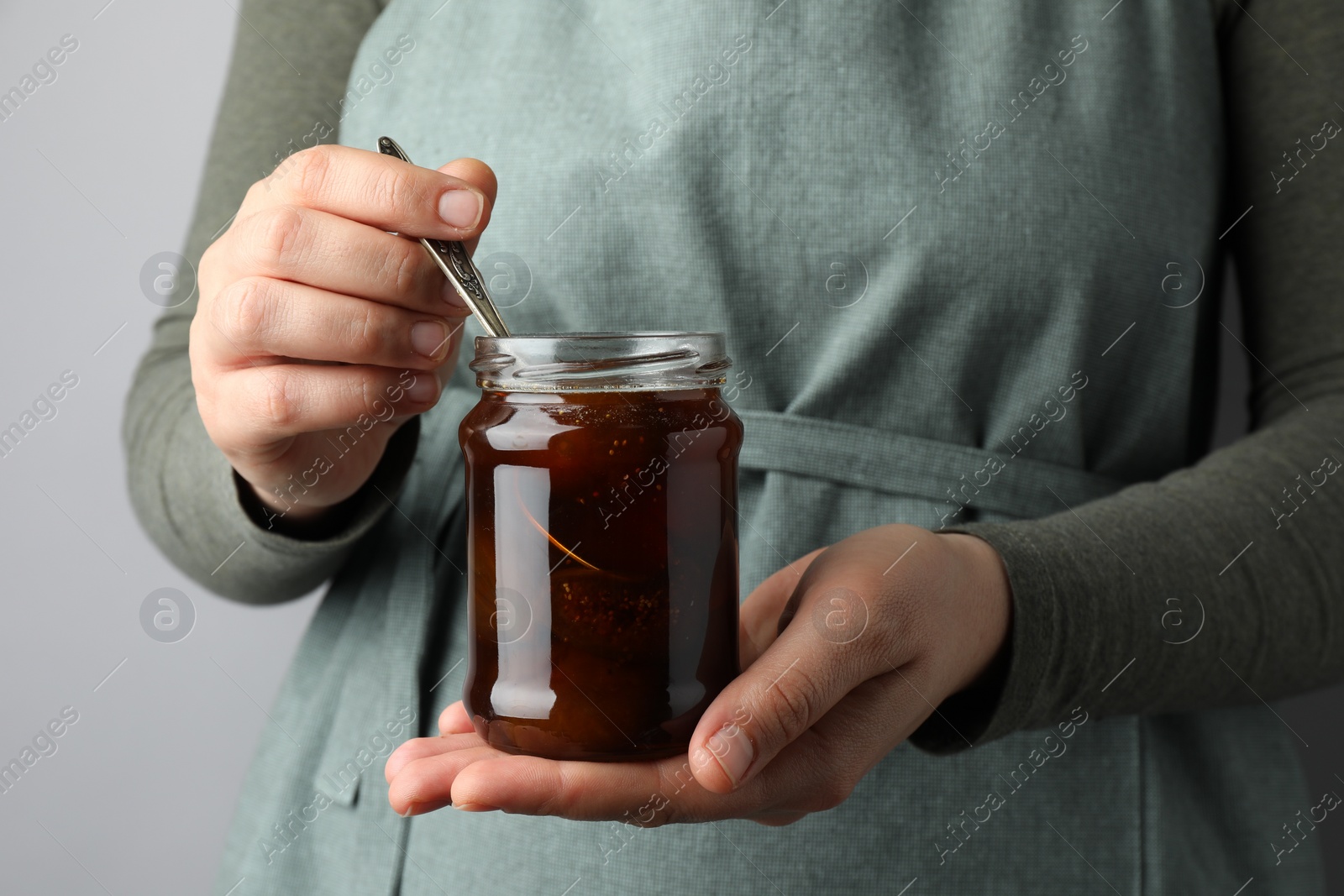 Photo of Woman holding jar of tasty sweet fig jam on grey background, closeup