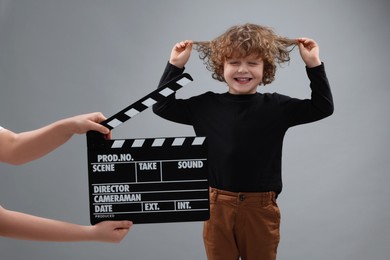 Photo of Happy cute boy performing while second assistant camera holding clapperboard on grey background. Little actor