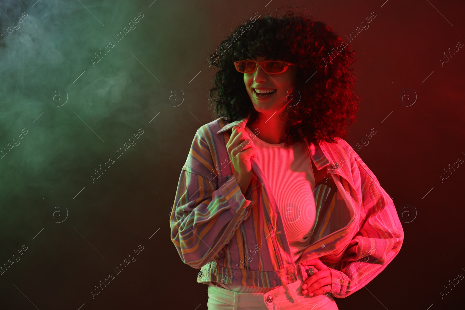 Photo of Beautiful young woman in sunglasses posing on color background in neon lights and smoke. Space for text