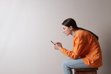 Photo of Young woman with bad posture using smartphone while sitting on stool against light grey background, space for text