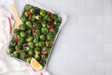 Delicious roasted Brussels sprouts, bacon and lemon in baking dish on white tiled table, top view. Space for text