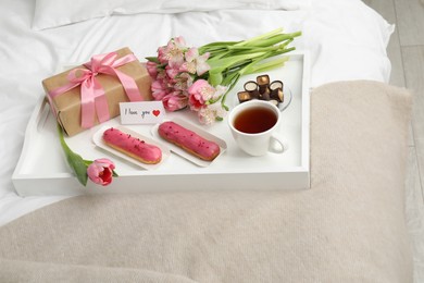 Photo of Tasty breakfast served in bed. Delicious desserts, tea, flowers, gift box and card with phrase I Love You on tray