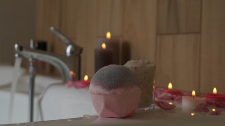 Colorful bath bomb, sea salt, flower petals and burning candles on white tub in bathroom