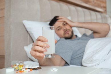 Man taking medicine for hangover in bed at home, focus on hand with glass