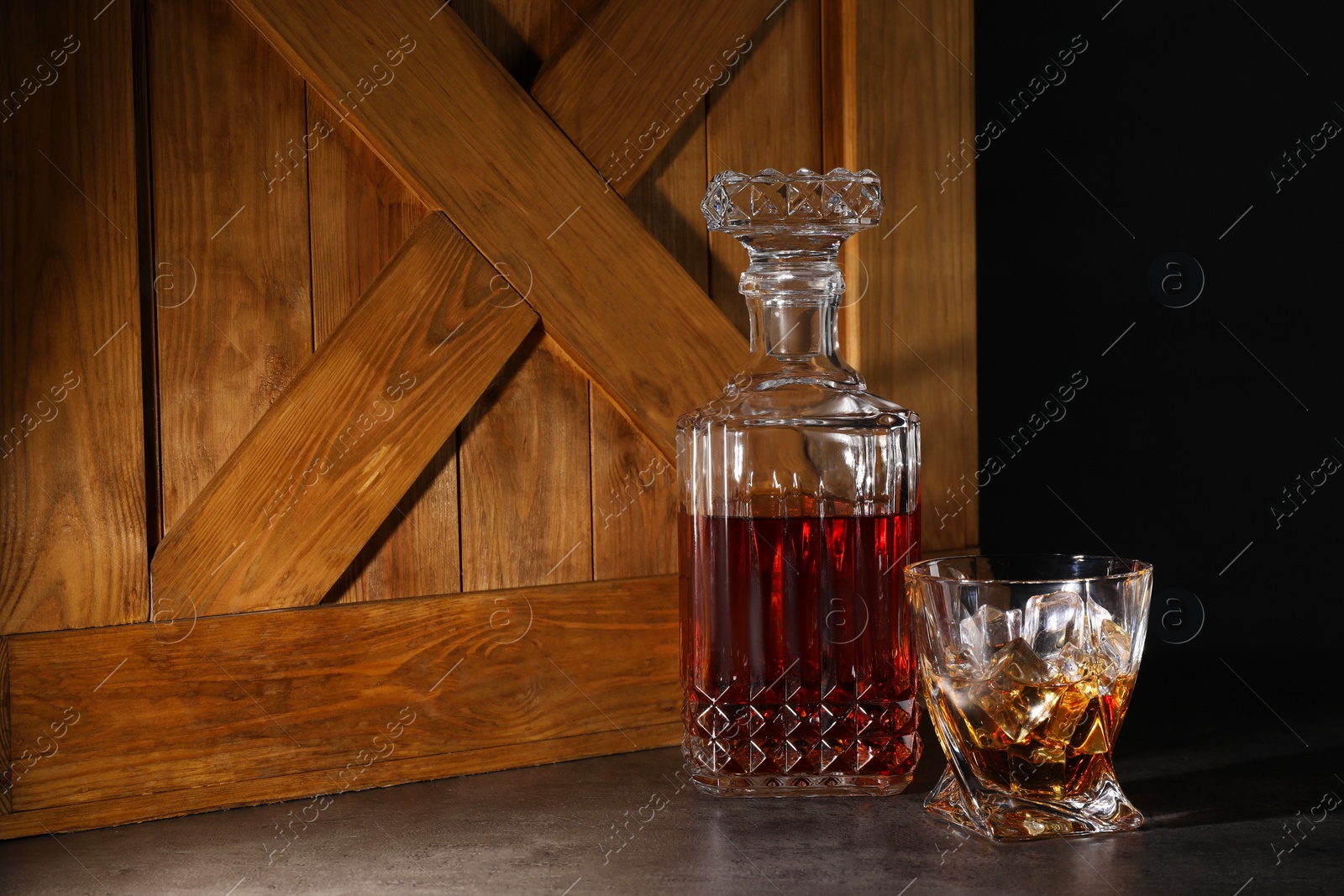 Photo of Whiskey in glass and bottle near wooden crate on dark table against black background