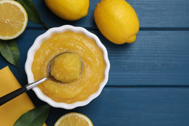 Photo of Delicious lemon curd in bowl, fresh citrus fruits and spoon on blue wooden table, flat lay. Space for text