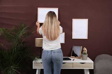Photo of Woman hanging empty frame on brown wall indoors, back view. Mockup for design