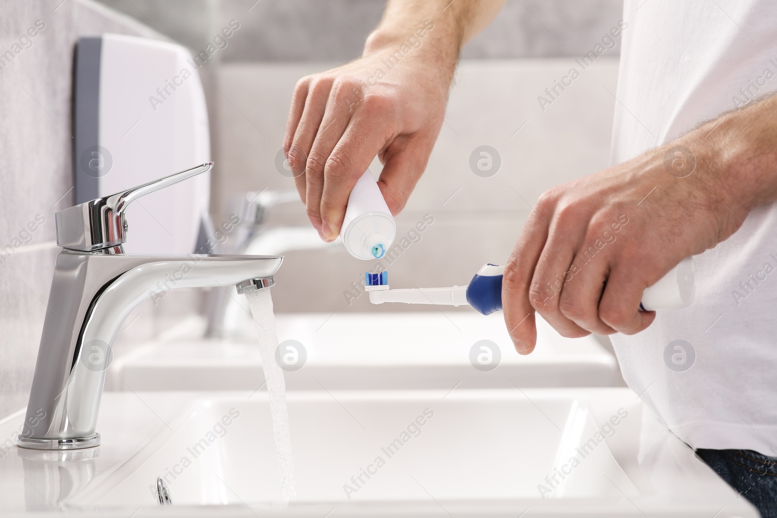 Photo of Man squeezing toothpaste from tube onto electric toothbrush above sink in bathroom, closeup