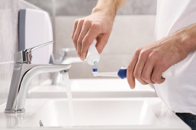 Man squeezing toothpaste from tube onto electric toothbrush above sink in bathroom, closeup