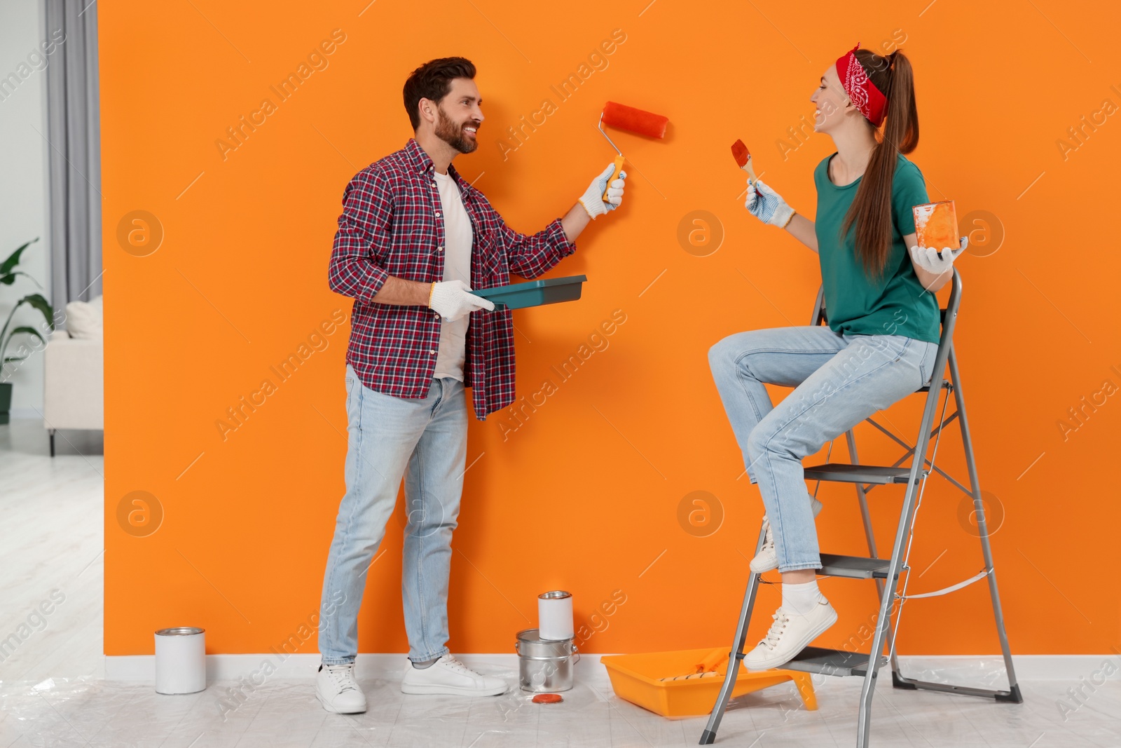 Photo of Man painting orange wall and happy woman holding can of dye with brush indoors. Interior design