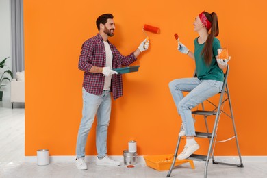 Photo of Man painting orange wall and happy woman holding can of dye with brush indoors. Interior design