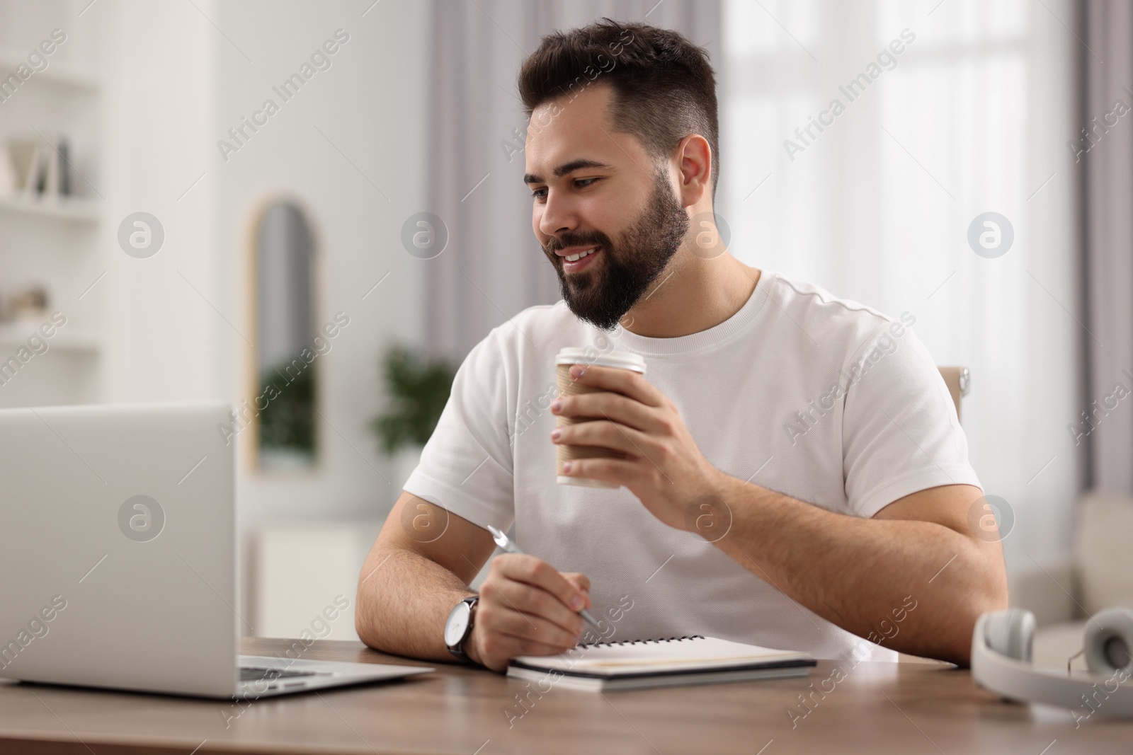 Photo of Young man with cup of coffee watching webinar at table in room