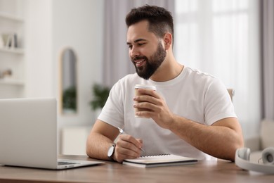 Young man with cup of coffee watching webinar at table in room