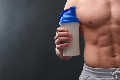 Young man with muscular body holding shaker of protein on dark grey background, closeup. Space for text