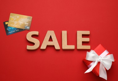 Word Sale made of wooden letters, credit cards and gift box on red background, flat lay