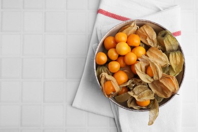 Ripe physalis fruits with calyxes in bowl on white tiled table, top view. Space for text