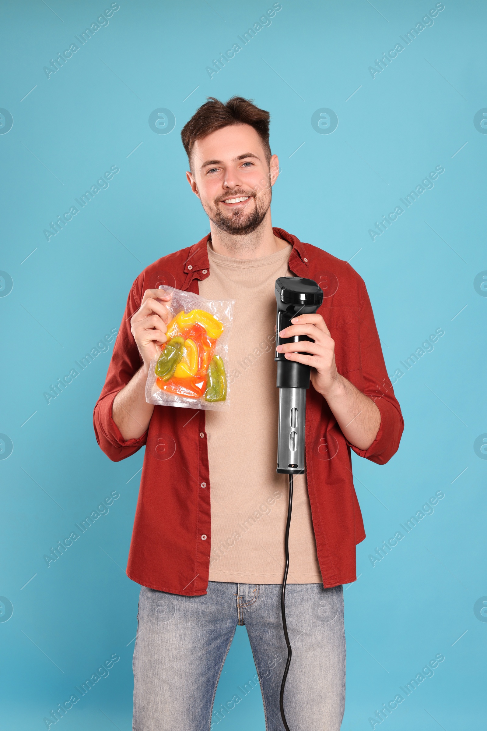 Photo of Smiling man holding sous vide cooker and vegetables in vacuum pack on light blue background