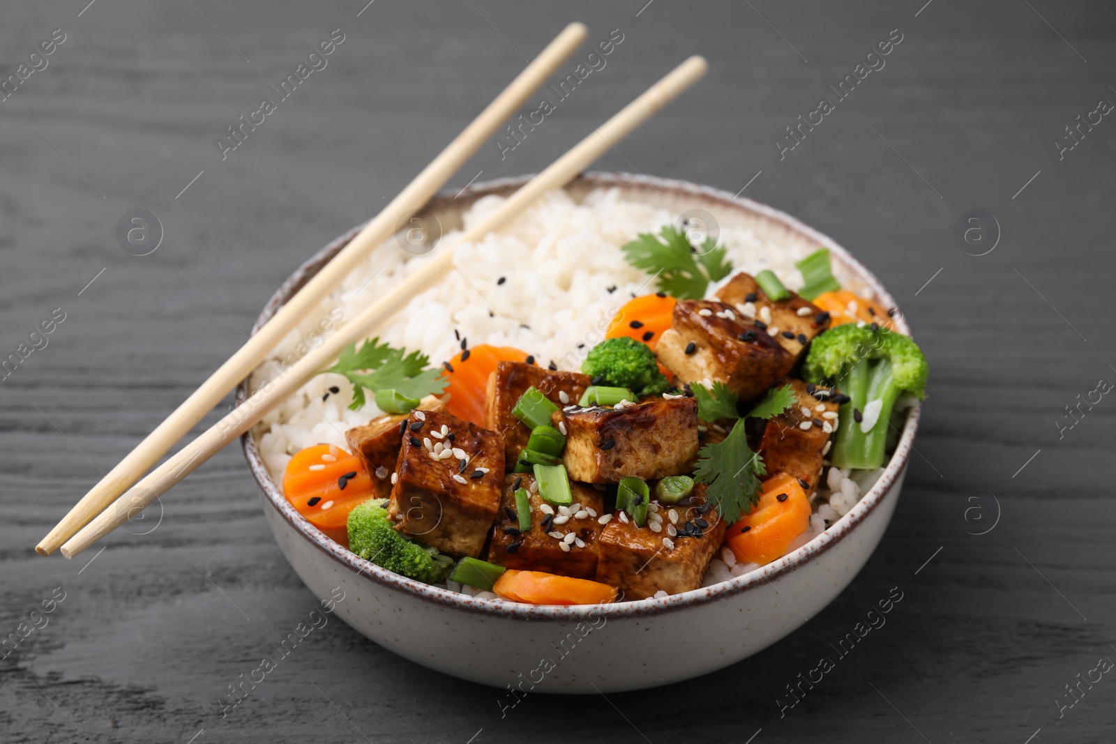 Photo of Bowl of rice with fried tofu, broccoli and carrots on grey wooden table, closeup