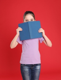 Cute little girl hiding behind book on color background. Reading activity