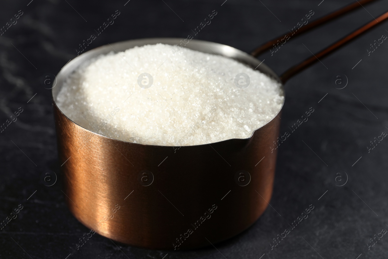 Photo of Metal measuring scoop of granulated sugar on black table, closeup