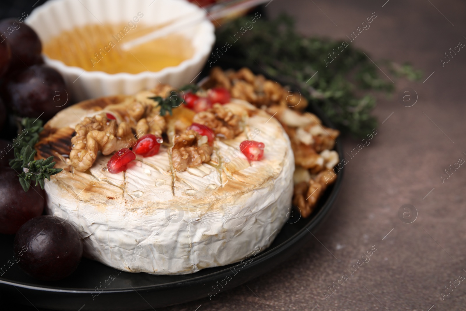 Photo of Plate with tasty baked camembert, honey, grapes, walnuts and pomegranate seeds on brown textured table, closeup