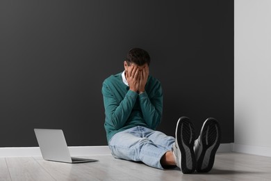 Photo of Upset man sitting on floor near laptop against black wall