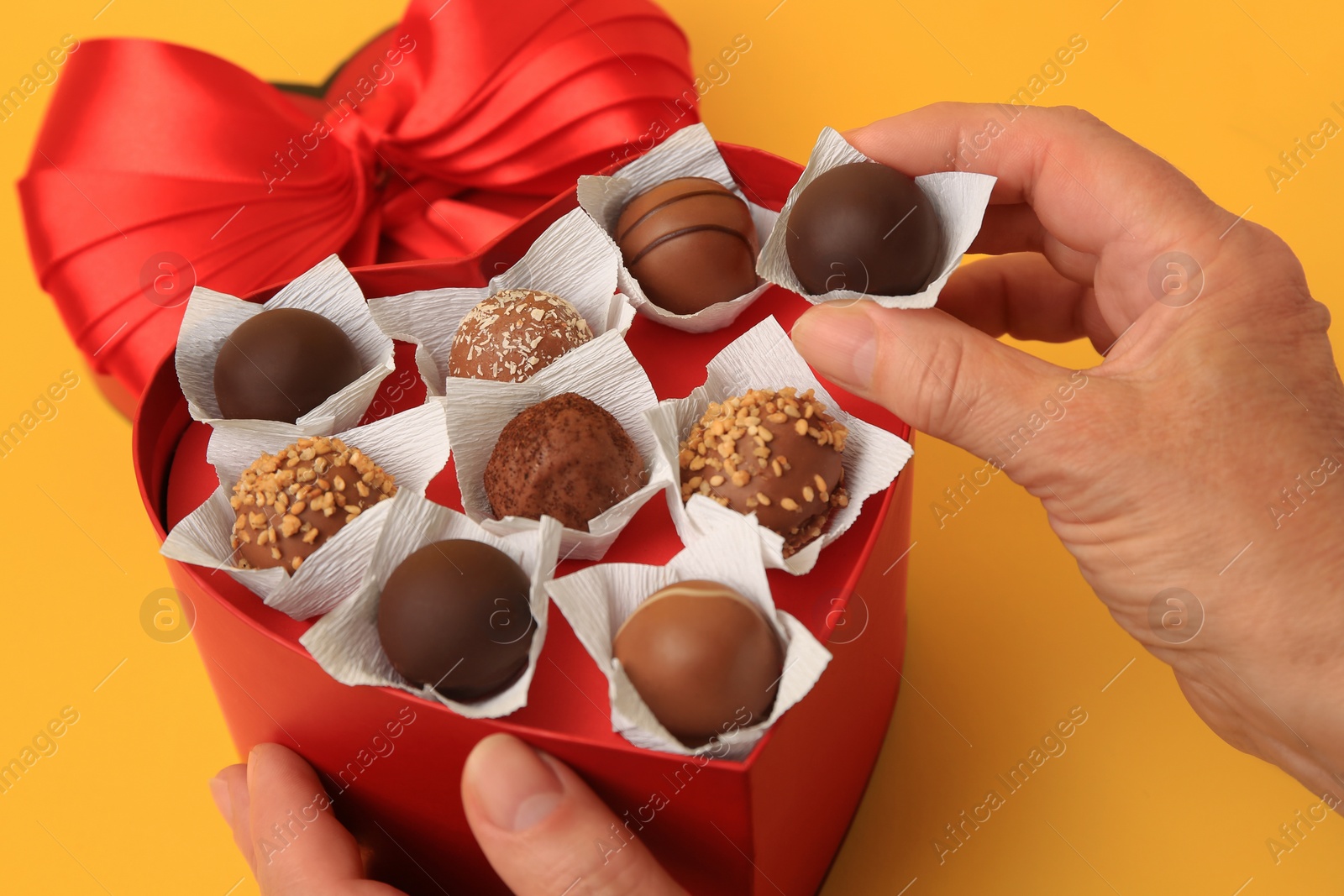 Photo of Woman taking delicious chocolate candy from heart shaped box on yellow background, closeup