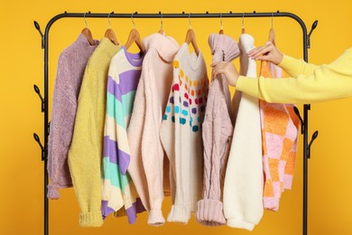 Photo of Woman taking stylish sweater from clothes rack against orange background, closeup