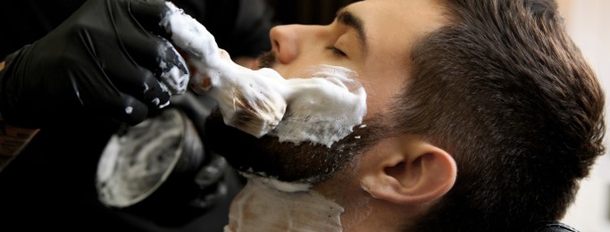 Image of Professional hairdresser applying shaving foam onto client's skin in barbershop, closeup. Banner design