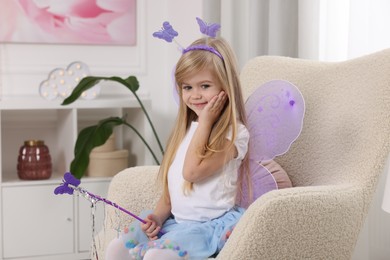 Photo of Cute little girl in fairy costume with violet wings and magic wand in armchair at home