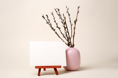 Photo of Vase with beautiful blooming willow branches and blank card on stand against beige background. Space for text