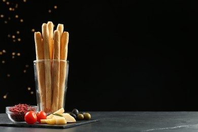 Photo of Fresh delicious grissini sticks in glass, red peppercorns, tomatoes, olives and cheese on dark table against black background, space for text