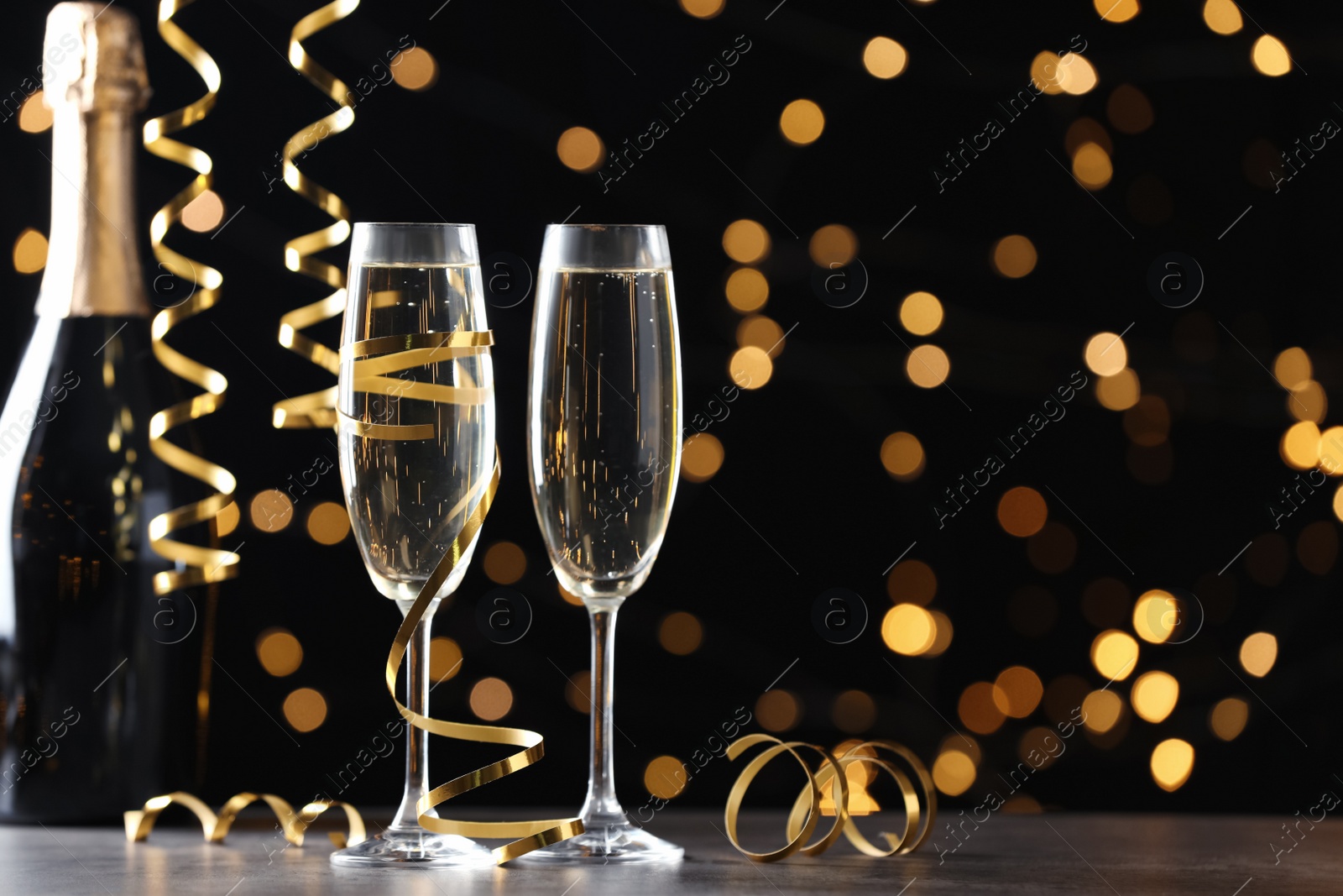 Photo of Glasses and bottle of champagne with serpentine streamers against blurred lights on black background. Space for text