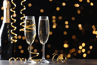 Glasses and bottle of champagne with serpentine streamers against blurred lights on black background. Space for text