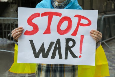 Photo of Woman wrapped in Ukrainian flag holding poster with words Stop War outdoors, closeup