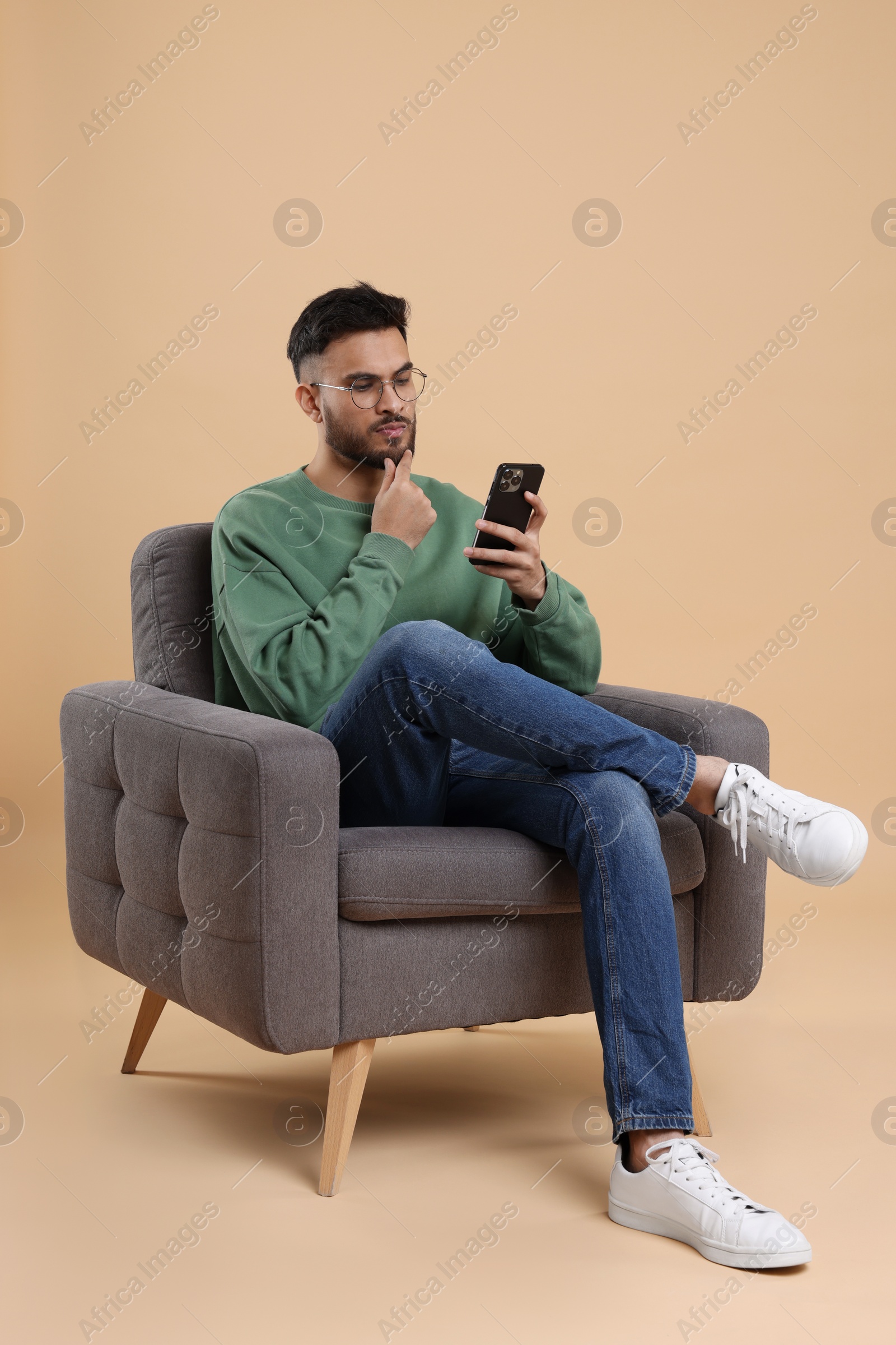 Photo of Handsome young man using smartphone in armchair on beige background