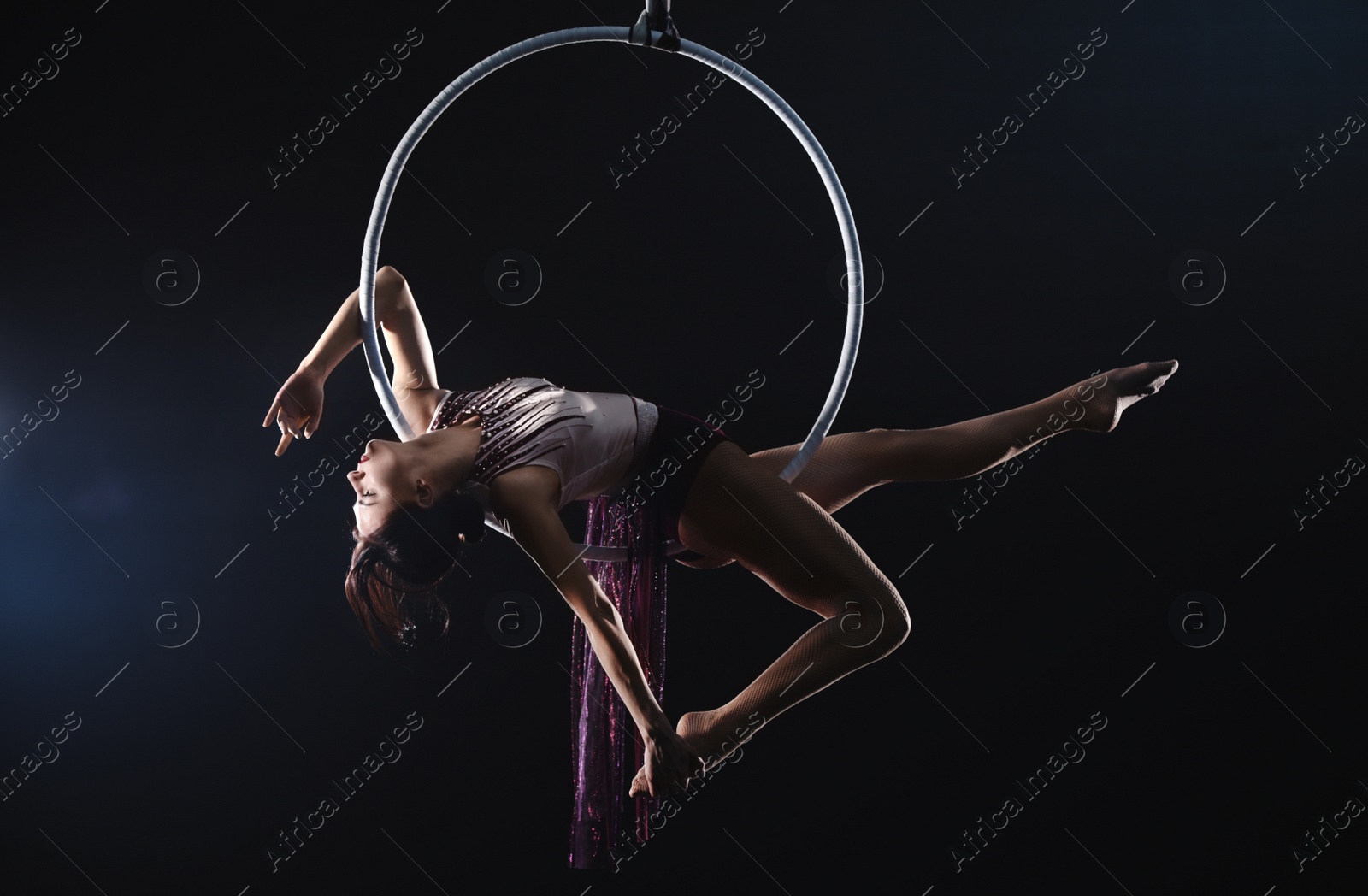 Photo of Young woman performing acrobatic element on aerial ring against dark background