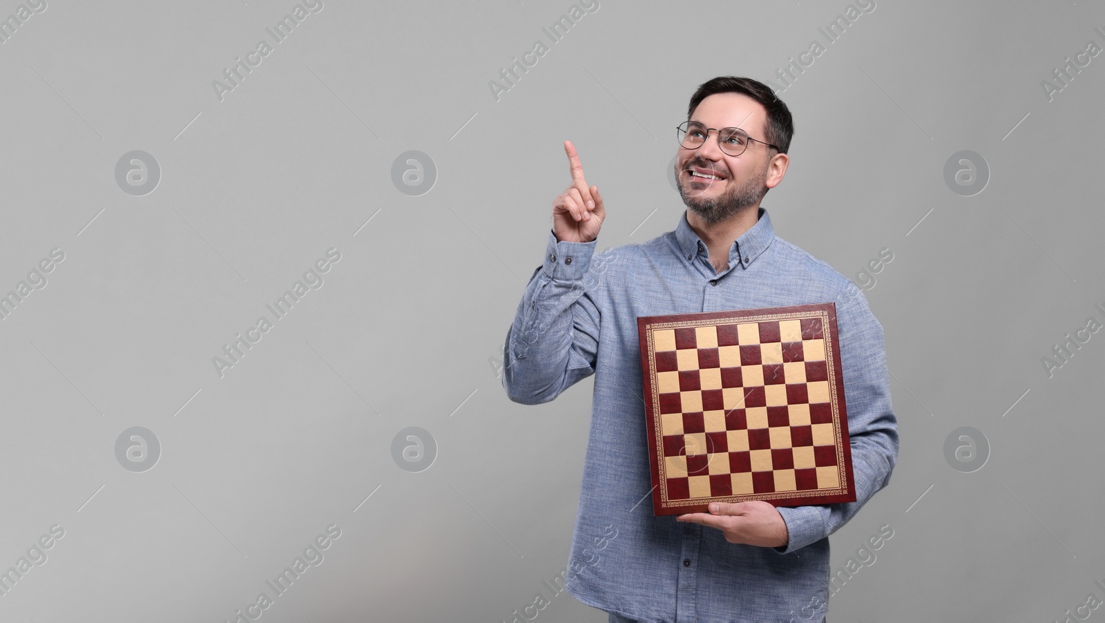 Photo of Smiling man holding chessboard and pointing up with finger on light grey background, space for text