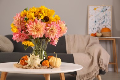 Beautiful bouquet with bright flowers and small pumpkins on white coffee table near sofa in room, space for text. Autumn atmosphere