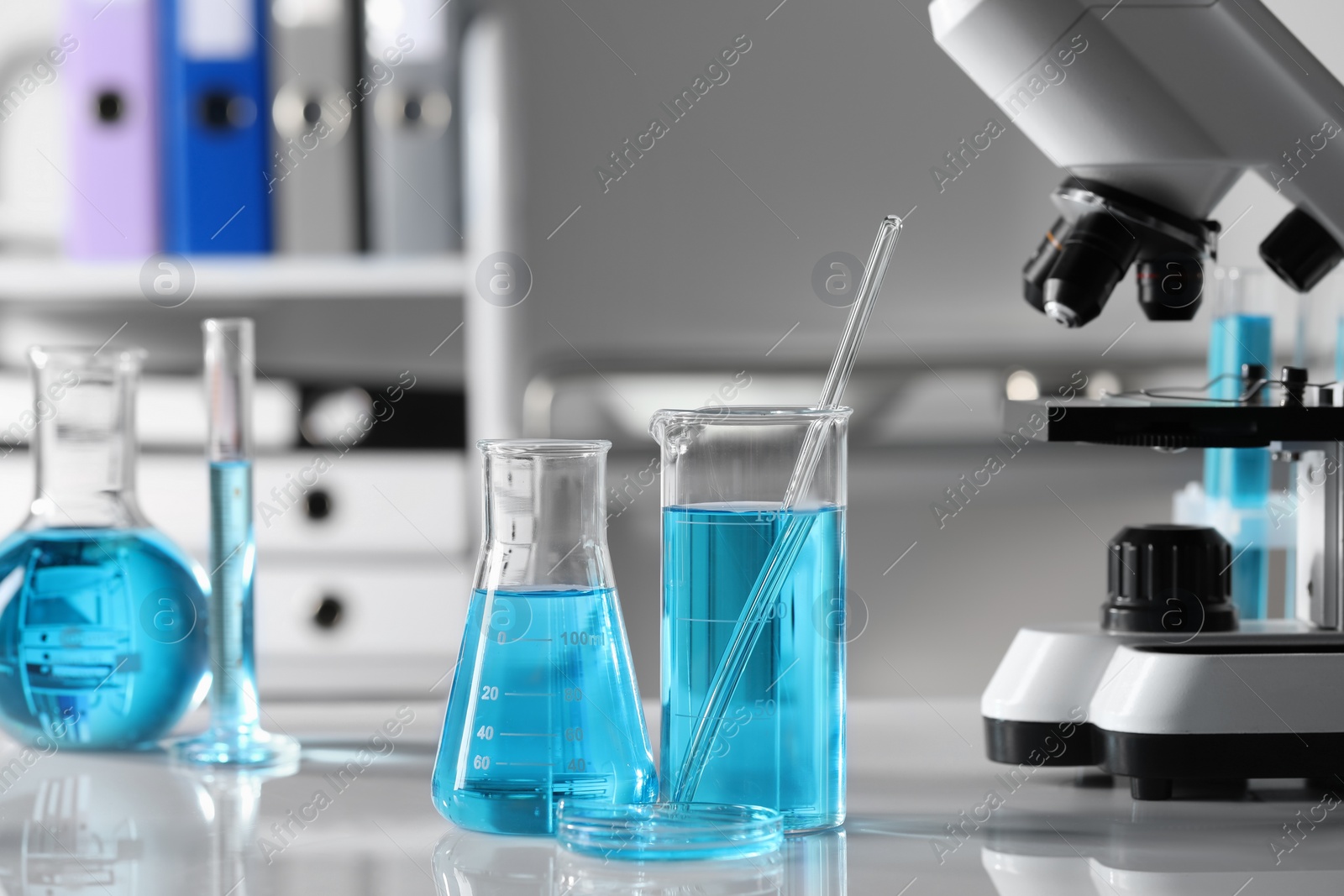 Photo of Different laboratory glassware with light blue liquid near microscope on table