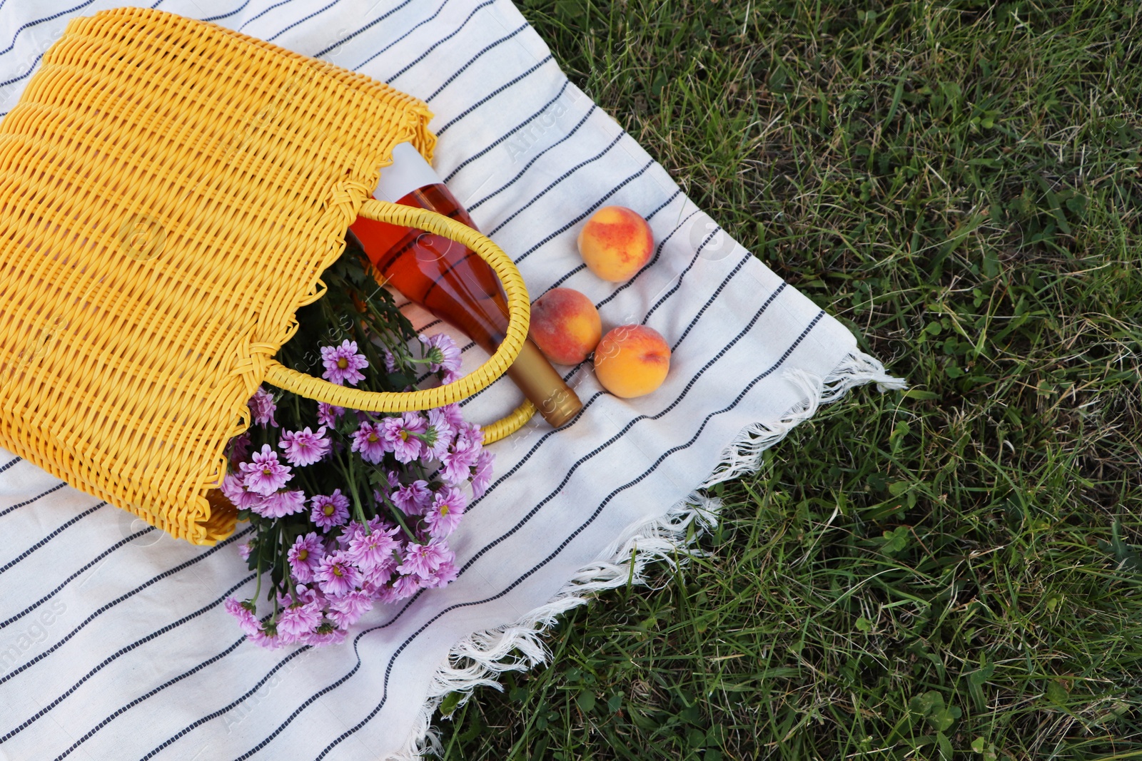 Photo of Yellow wicker bag with beautiful flowers, bottle of wine and peaches on picnic blanket outdoors
