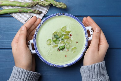 Woman with bowl of delicious asparagus soup at blue wooden table, top view