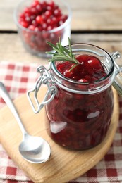 Photo of Fresh cranberry sauce in glass jar, rosemary and spoon on table